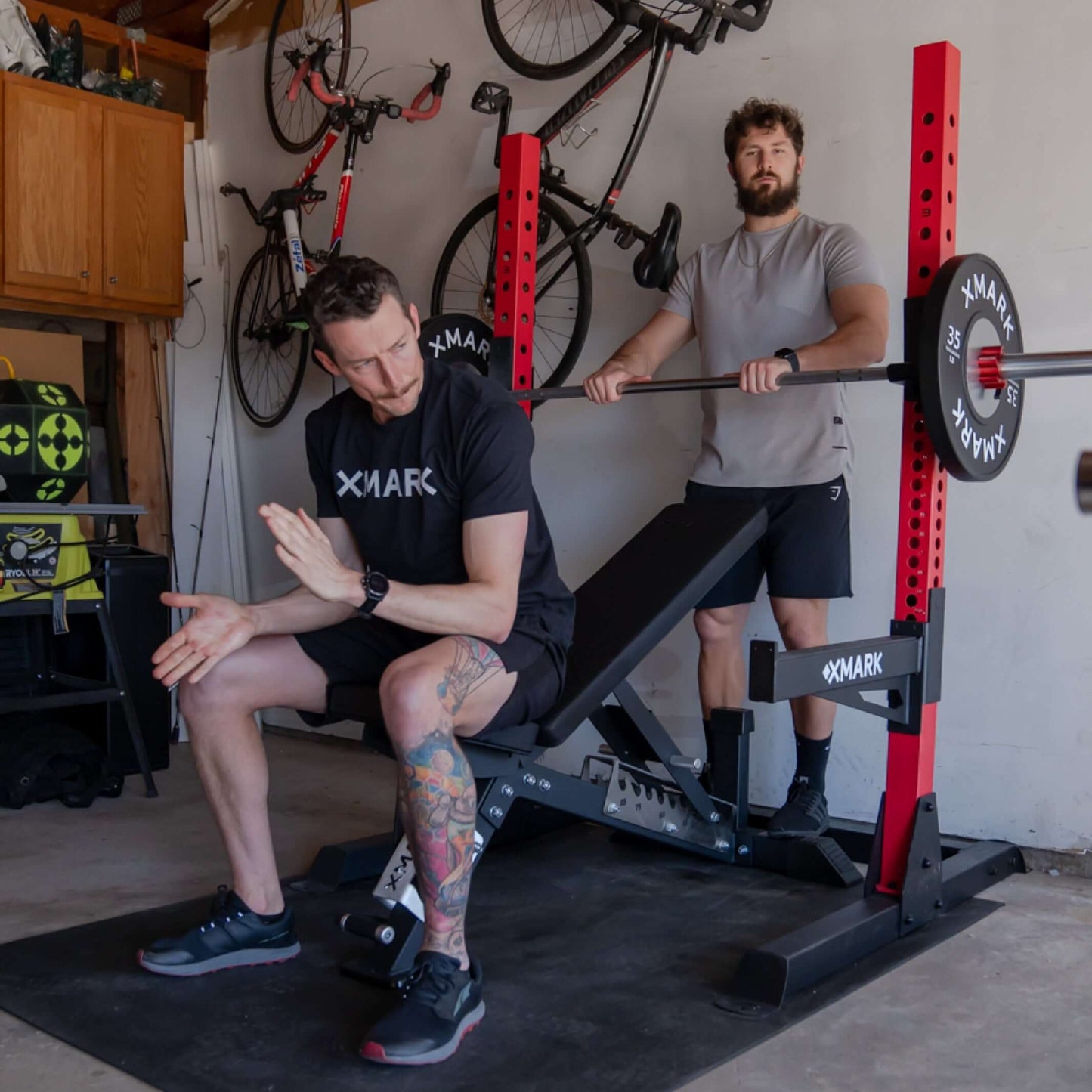 Two men using a Black Hawk Adjustable Weight Bench in a garage gym setting, one is demonstrating an exercise while the other observes.